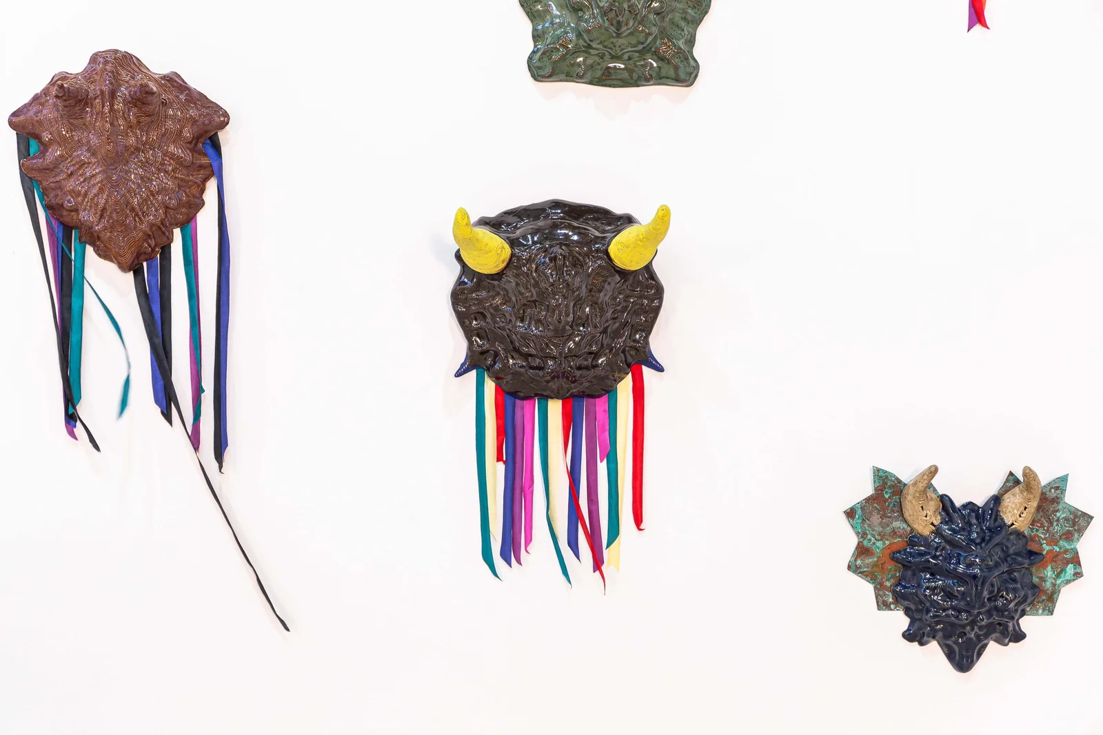 Protocolos de Emergencia (Emergency Protocols), installation view, three glazed ceramic masks with horns and long teal, yellow, and purple ribbons on a white wall