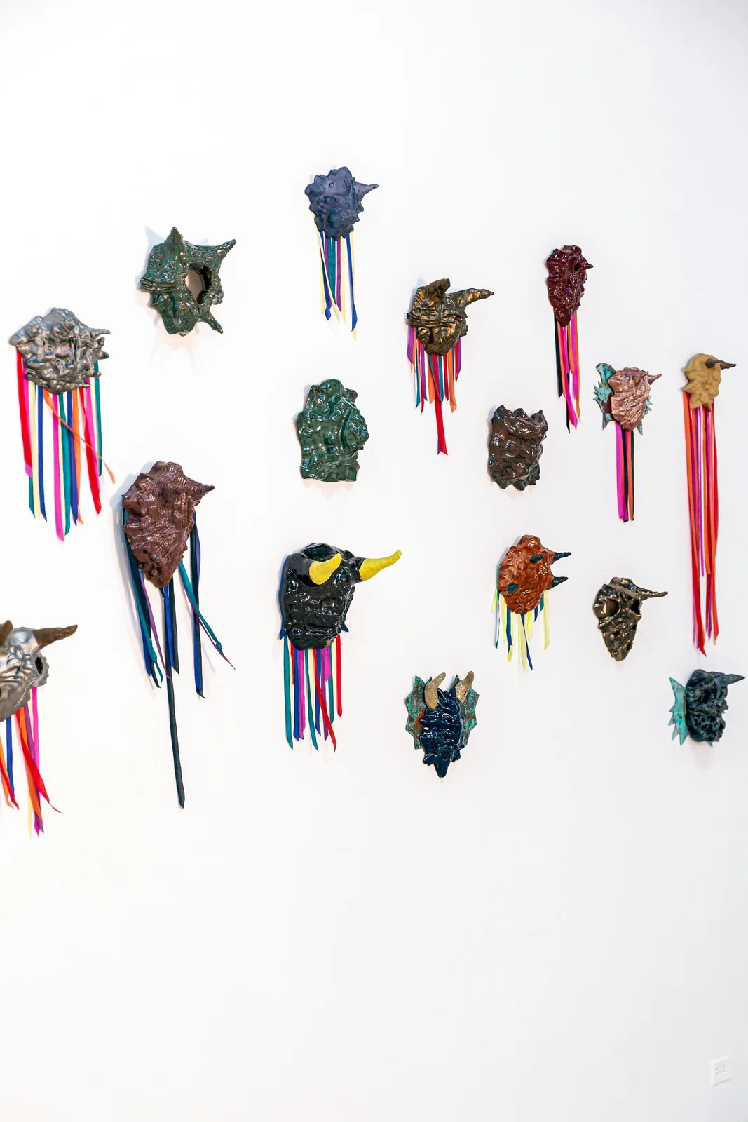 Protocolos de Emergencia (Emergency Protocols), installation view, wide shot of many colorful glazed masks with ribbon fringe arranged across a white gallery wall