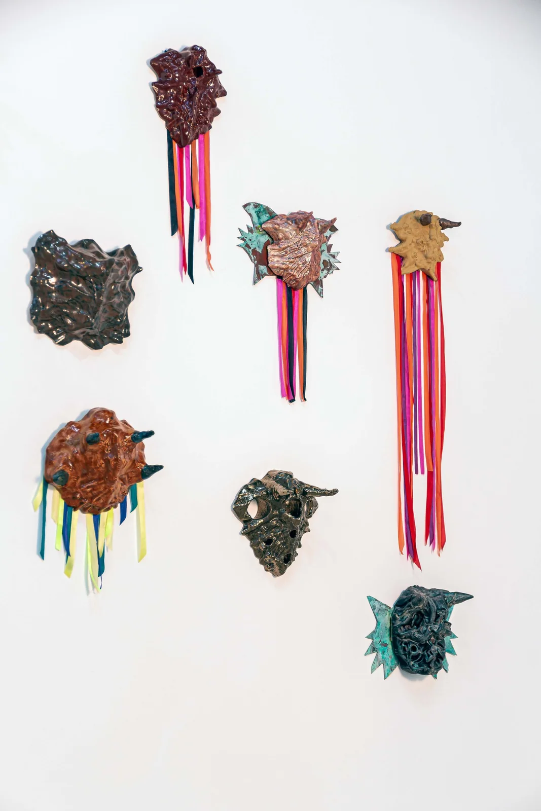 Protocolos de Emergencia (Emergency Protocols), installation view, seven abstract wall-mounted sculptures in brown and green glazes with pink, orange, and blue ribbons