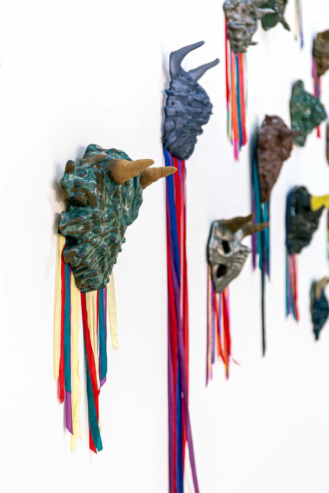 Protocolos de Emergencia (Emergency Protocols), installation view, row of teal horned mask sculptures with red and yellow ribbons receding along a white wall, shallow depth of field