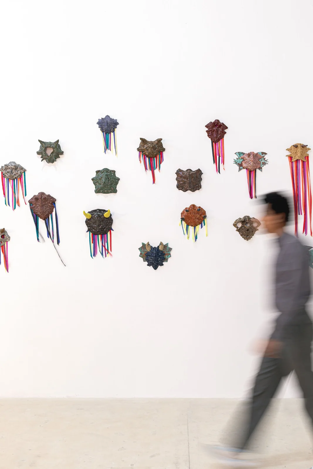 Protocolos de Emergencia (Emergency Protocols), installation view, scattered mask sculptures with ribbons on a white wall, motion-blurred figure crossing the frame