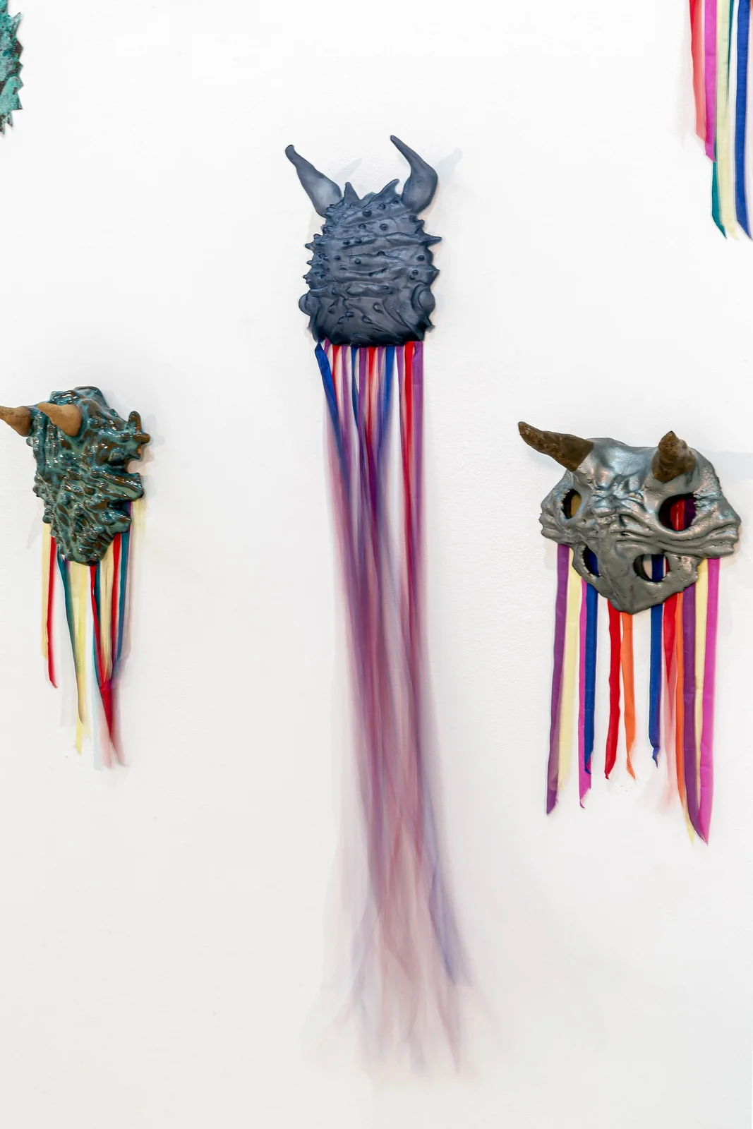 Protocolos de Emergencia (Emergency Protocols), installation view, three wall-mounted horned masks in teal, dark blue, and silver with long colored and sheer ribbons
